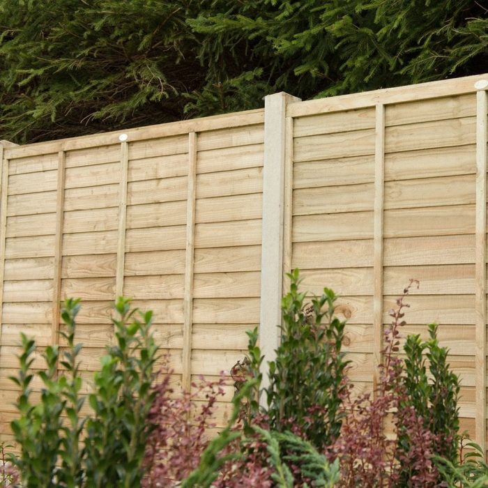 Wooden fence with vertical and horizontal panels, surrounded by green and purple shrubs. Tall evergreen trees form a lush backdrop, conveying tranquility.