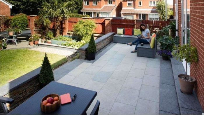 Modern patio with a woman reading on a bench, colourful cushions, and lush plants. A table holds a fruit bowl and notebook. Red-brick houses in the background.