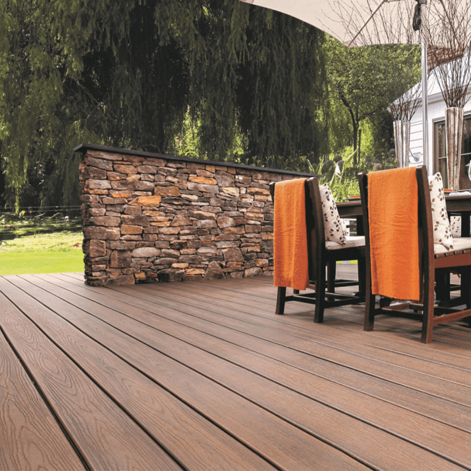 Spacious wooden deck with a stone wall backdrop, orange towels draping over patio chairs. Greenery surrounds, creating a lush, serene atmosphere.